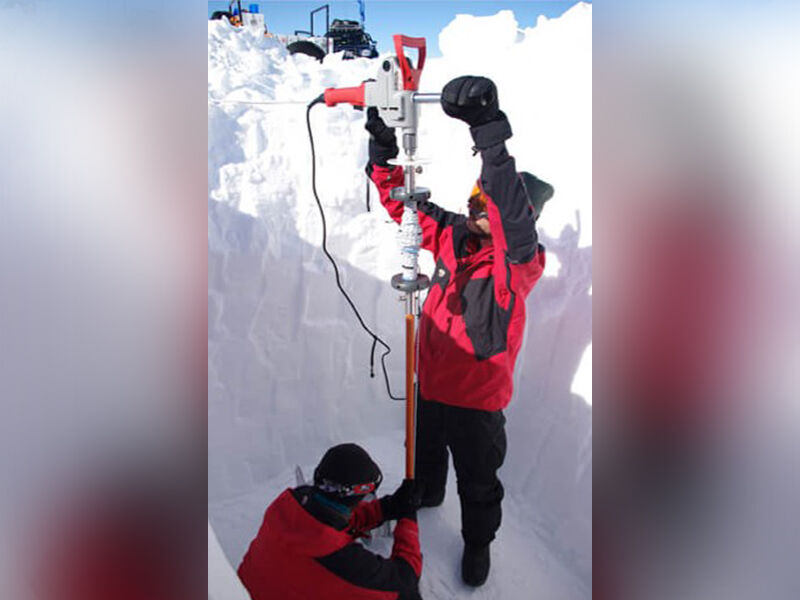 Two scientists in the snow with ice drilling equipment, preparing to drill into the snowpack beneath them. The orange tubing is visible on lower half of the equipment. 